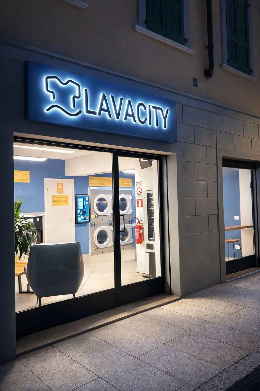 L'ingresso di LAVACITY in Via Gianbattista Moroni 150 a Bergamo di sera, con l'insegna accesa e le lavatrici visibili dalla vetrina